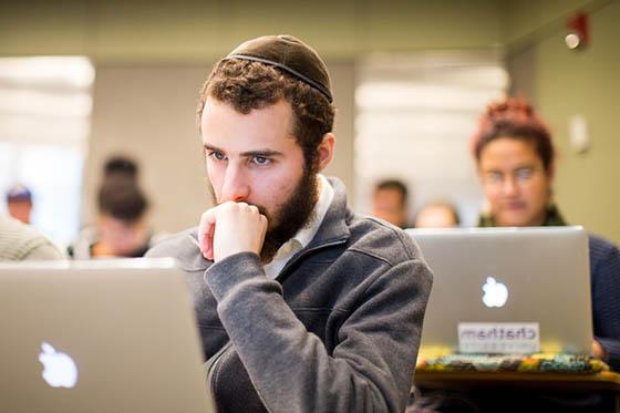 一个戴圆顶小帽的男学生的照片, sitting in a Chatham University lecture hall and looking at his computer